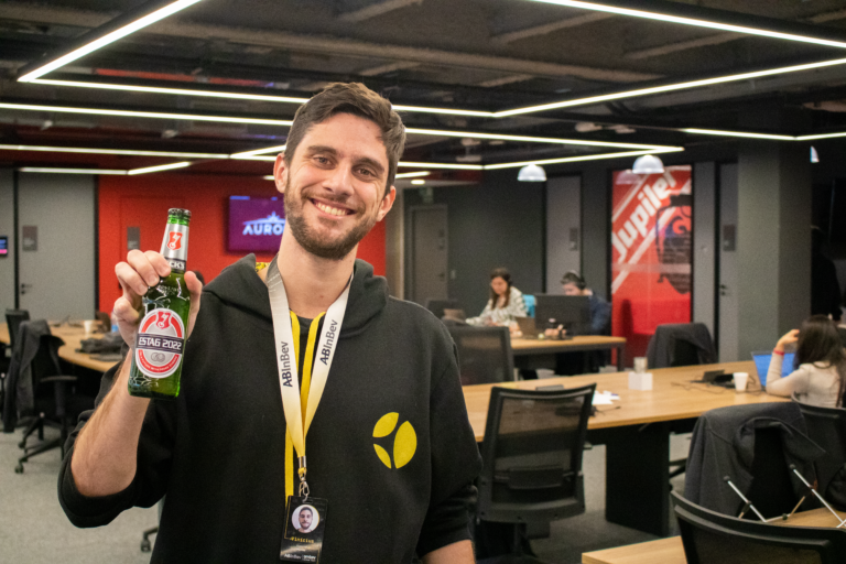 Homem jovem no primeiro plano, segurando uma cerveja, ao fundo o escrit[orio.