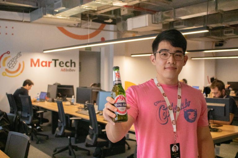 Homo jovem no primeiro plano, segurando uma cerveja, ao fundo o escrit[orio.