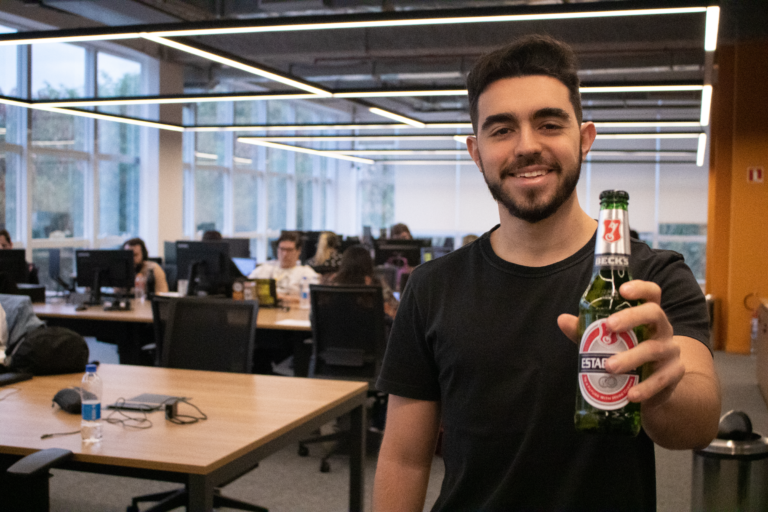 Homo jovem no primeiro plano, segurando uma cerveja, ao fundo o escrit[orio.