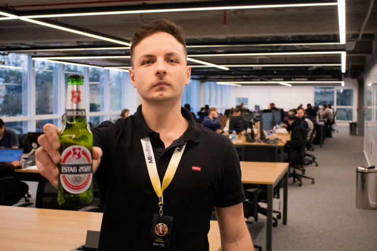 Homem jovem no primeiro plano, segurando uma cerveja, ao fundo o escrit[orio.